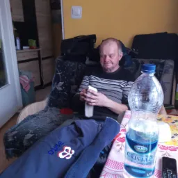 Mężczyzna siedzi w fotelu, odkręcając butelkę wody. Na stole stoi duża butelka wody mineralnej Cristal i stary telefon komórkowy. W tle widać szafkę i walizkę.