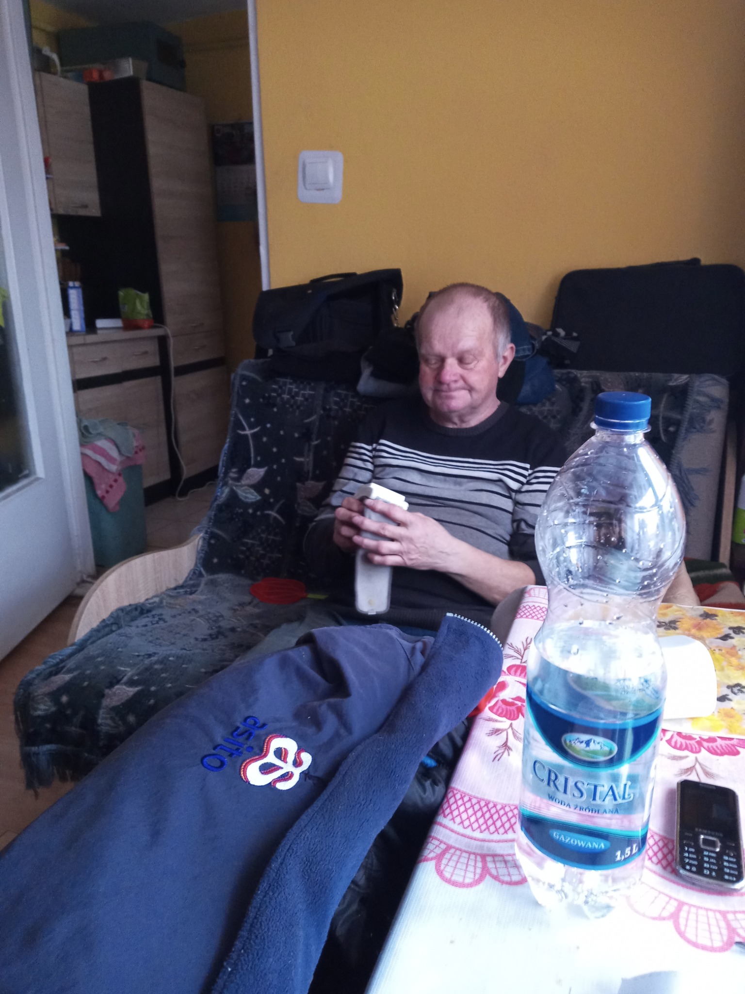 Mężczyzna siedzi w fotelu, odkręcając butelkę wody. Na stole stoi duża butelka wody mineralnej Cristal i stary telefon komórkowy. W tle widać szafkę i walizkę.