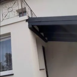 Fragment elewacji budynku z balkonem z ozdobną, metalową balustradą i zadaszeniem nad wejściem. Świeżo otynkowana ściana w jasnym odcieniu.