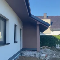 Elewacja domu w trakcie prac wykończeniowych: biała ściana z szarym cokołem, brązowy fragment elewacji pod dachem, widoczny system rynnowy, teren wokół domu w trakcie zagospodarowania.
