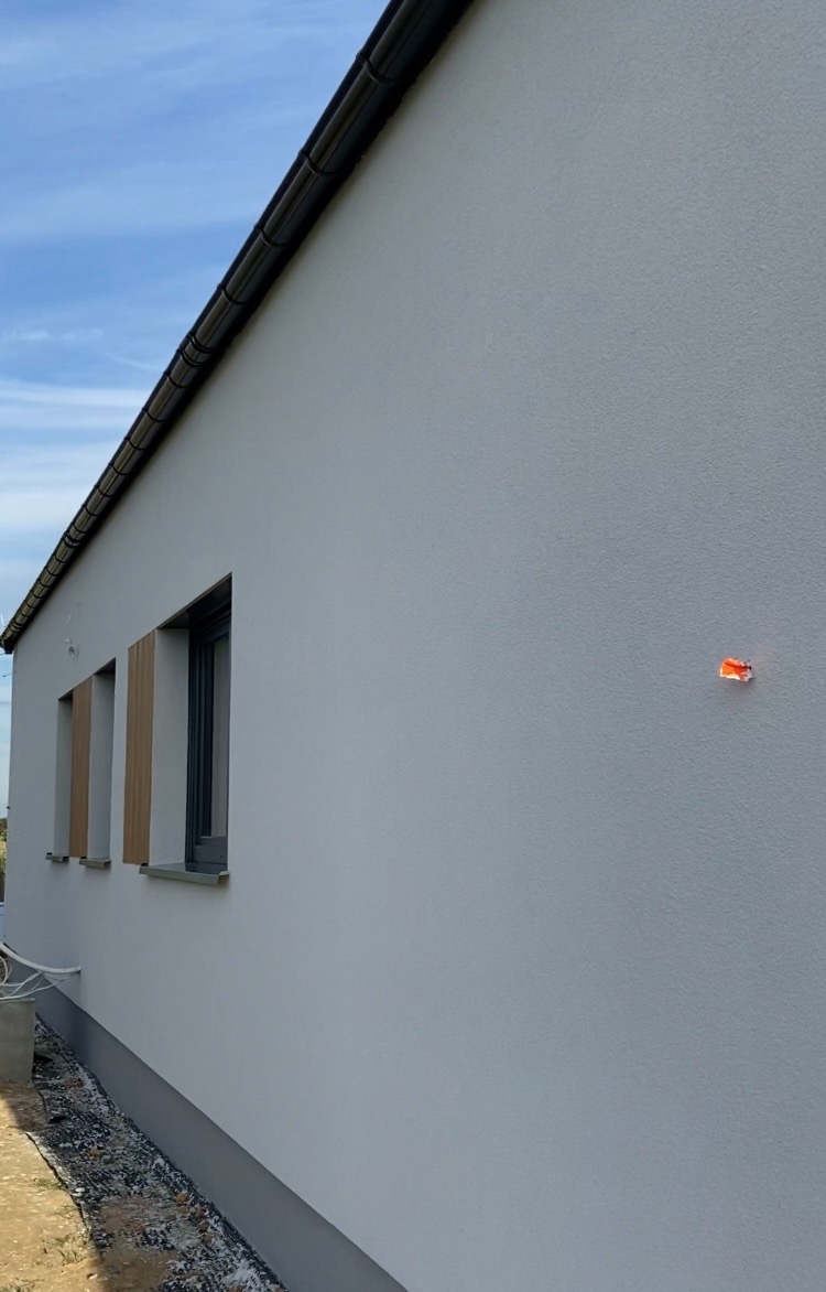 Świeżo otynkowana elewacja domu jednorodzinnego w jasnym odcieniu, widoczne trzy okna z drewnianymi okiennicami, czarne rynny i fragment nieba.