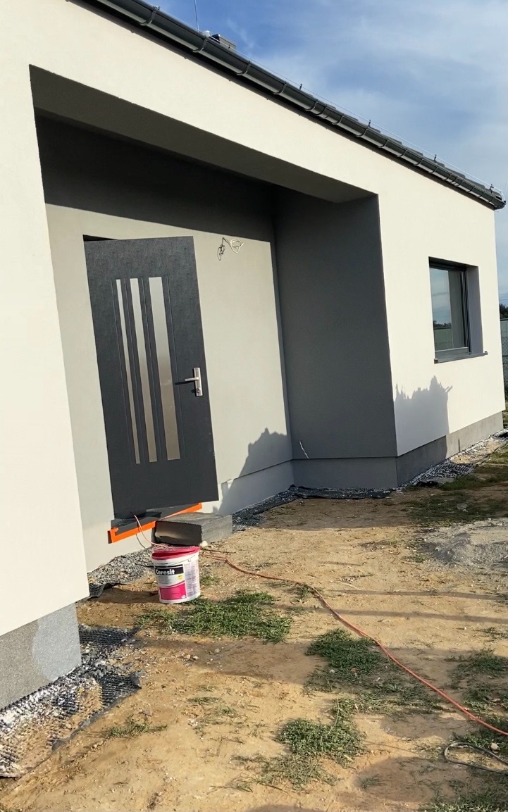 Nowoczesny dom jednorodzinny po tynkowaniu elewacji na szaro i biało, widoczny fragment ocieplenia przy drzwiach wejściowych oraz wiadro z farbą marki Ceresit.