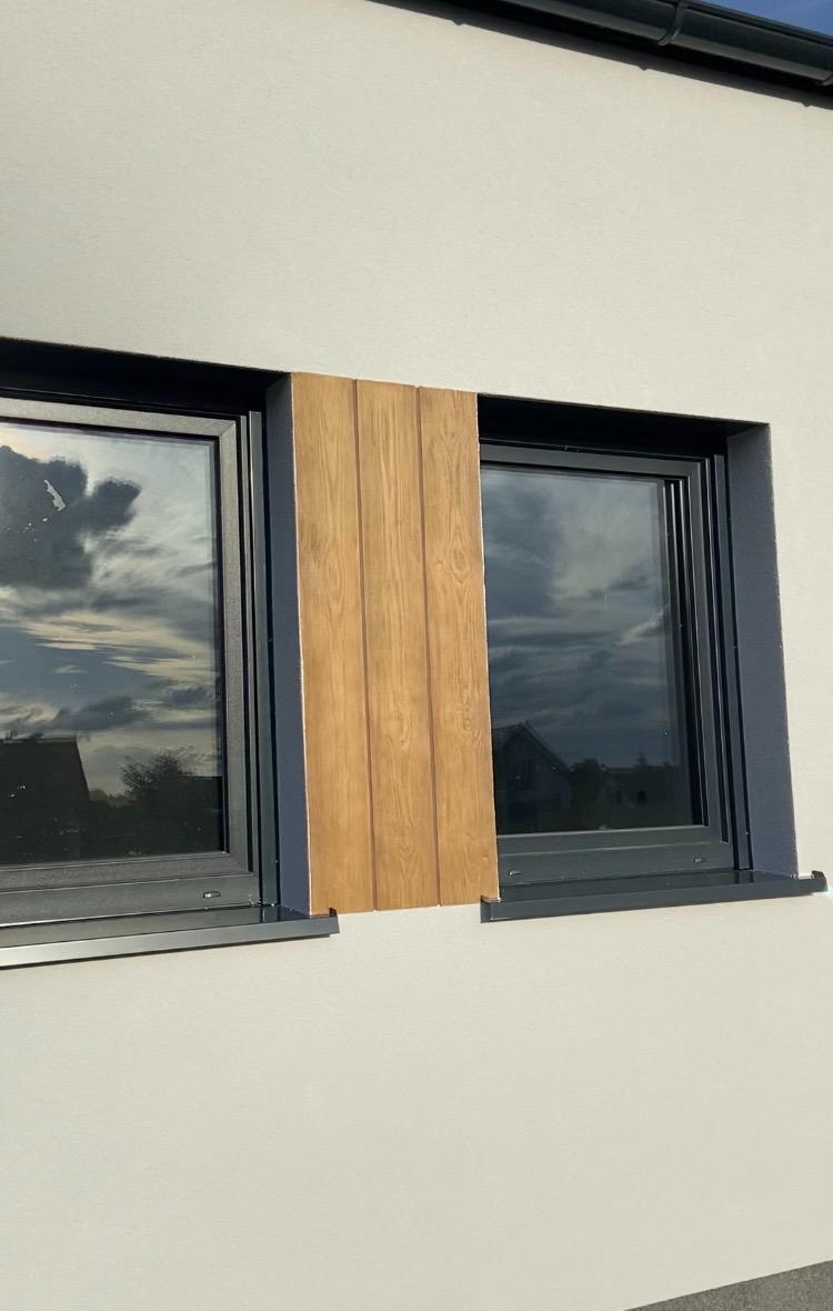 Fragment ocieplonej elewacji z okładziną drewnopodobną między dwoma oknami, widoczna faktura tynku strukturalnego i odbicie chmur w szybach.