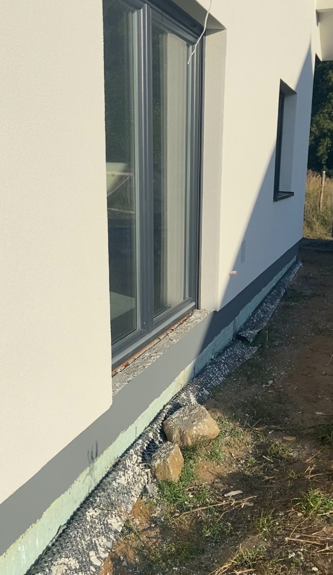 Fragment elewacji budynku z oknem i widocznym ociepleniem fundamentów oraz fragmentem trawnika z dwoma kamieniami.