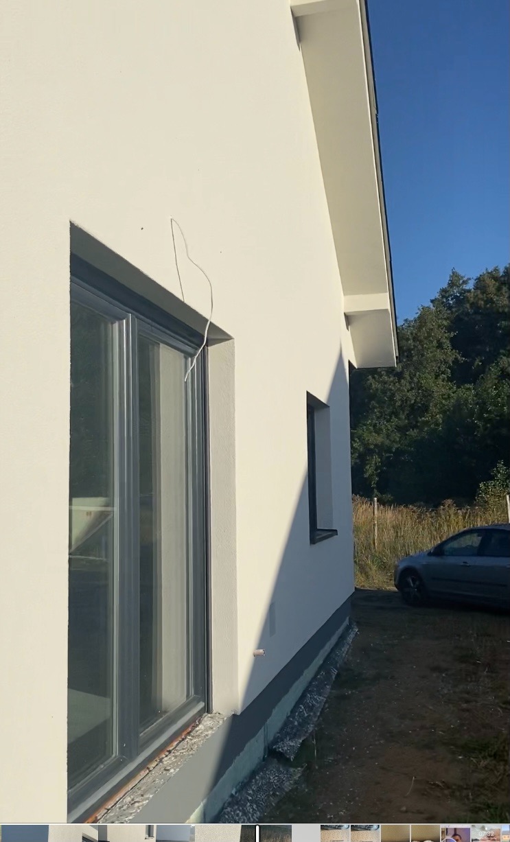 Ściana domu z oknem po tynkowaniu, widoczny fragment wystającego przewodu elektrycznego, szara podmurówka i cień padający na elewację.