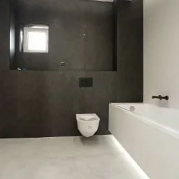 Minimalistyczna łazienka z szarymi płytkami imitującymi kamień, podświetlana wanna i wisząca toaleta, małe okno w głębi.