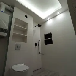 Nowoczesna łazienka z białymi płytkami, czarną armaturą, podświetlanym sufitem i drewnopodobną podłogą, widoczny wiszący sedes i wnęka na kosmetyki.