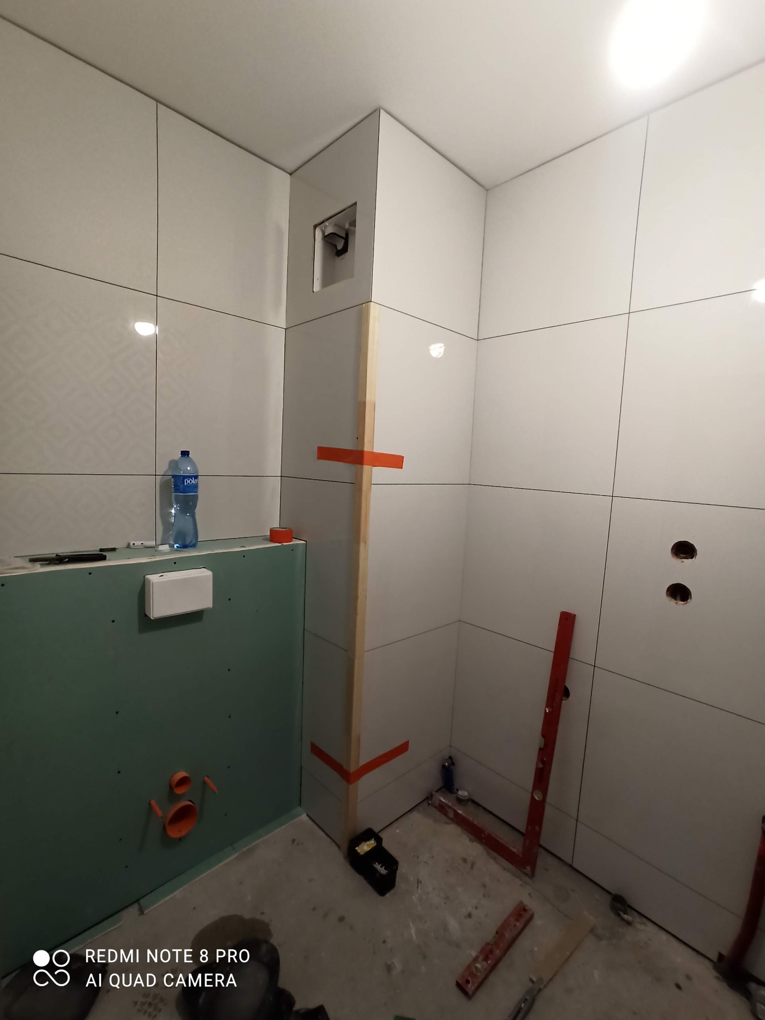 Łazienka w trakcie remontu: białe płytki z delikatnym wzorem na ścianach, zielona płyta gipsowo-kartonowa z instalacją hydrauliczną, butelka wody, poziomica i inne narzędzia na podłodze, widoczne...