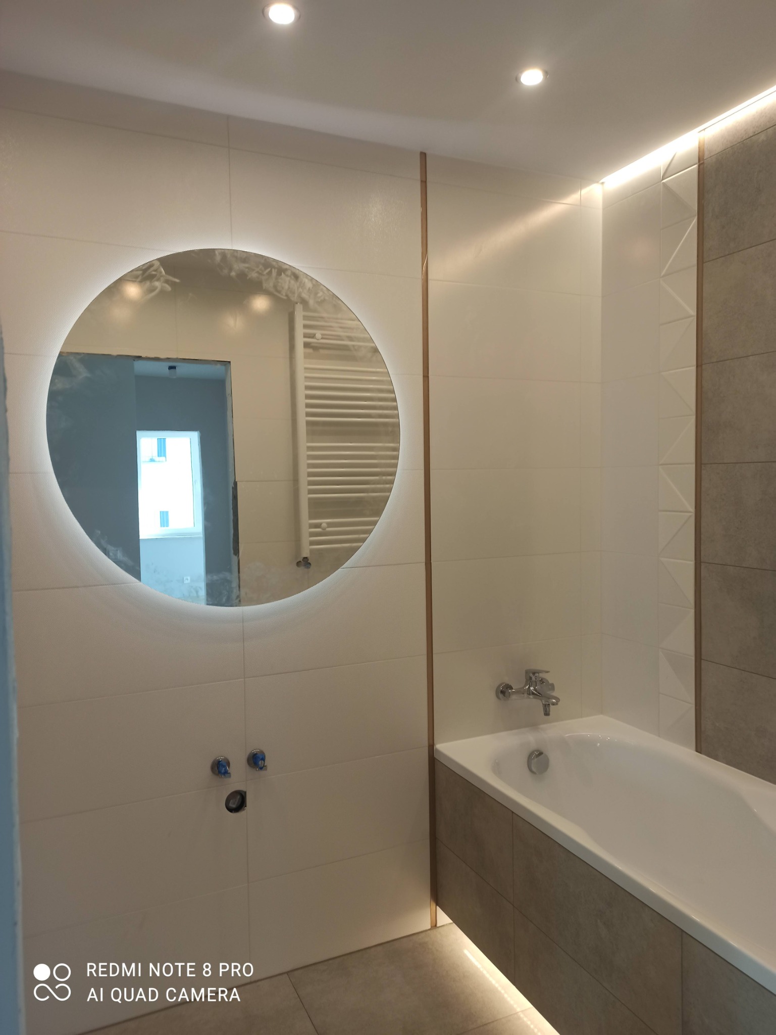 Nowoczesna łazienka z podświetlanym okrągłym lustrem, biało-szare płytki z dekoracyjnymi listwami, wanna z obudową z płytek, widoczne przyłącza wodne i grzejnik drabinkowy w odbiciu lustra.