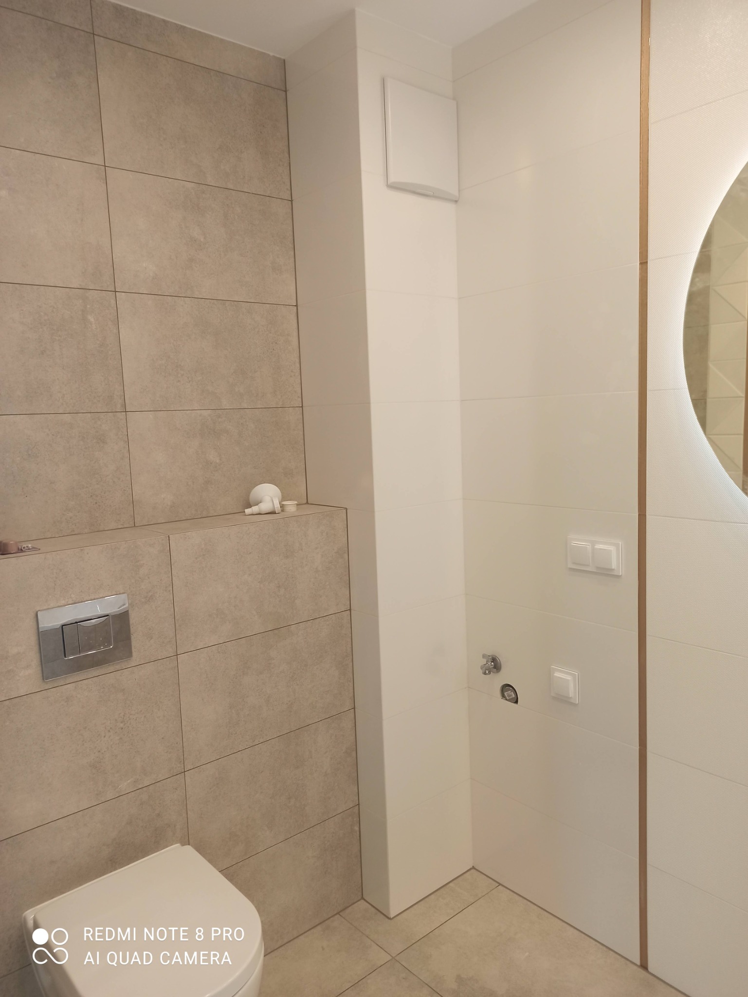 Nowoczesna łazienka z beżowymi i białymi płytkami, wisząca toaleta z chromowanym przyciskiem, okrągłe lustro z podświetleniem LED i wentylator na ścianie.