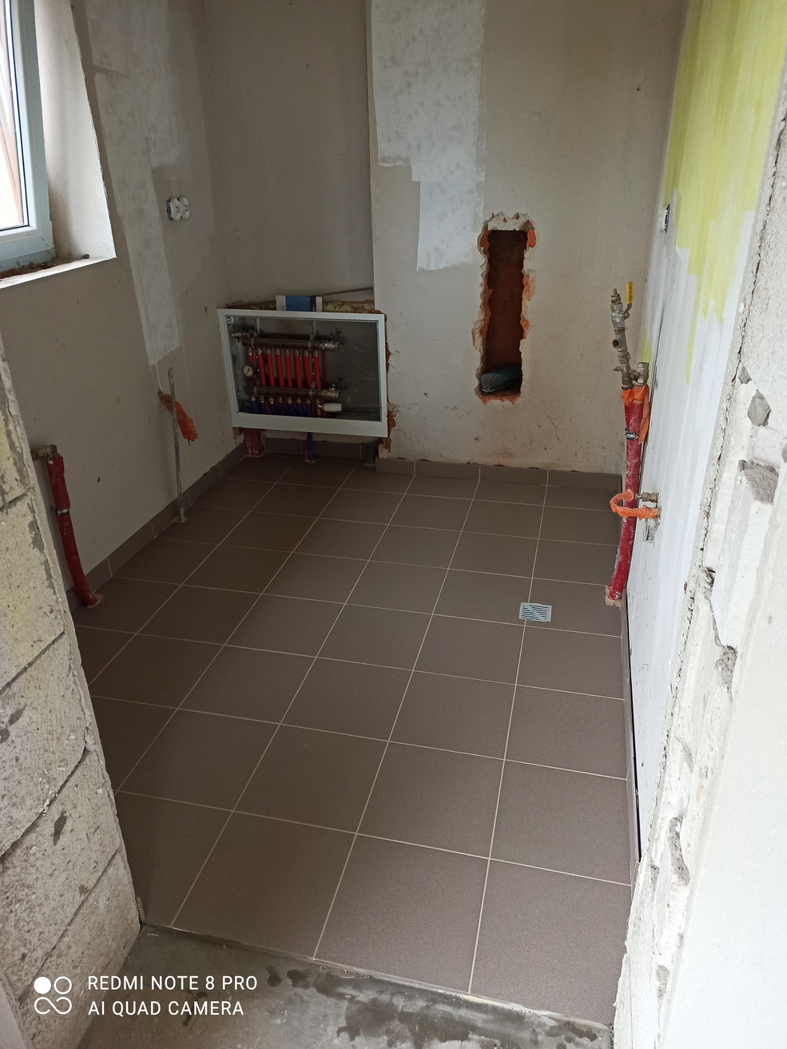 Łazienka w trakcie prac: ułożona brązowa terakota na podłodze, ściany częściowo przygotowane do malowania, widoczne rury instalacji grzewczej w ścianie i otwór w murze.