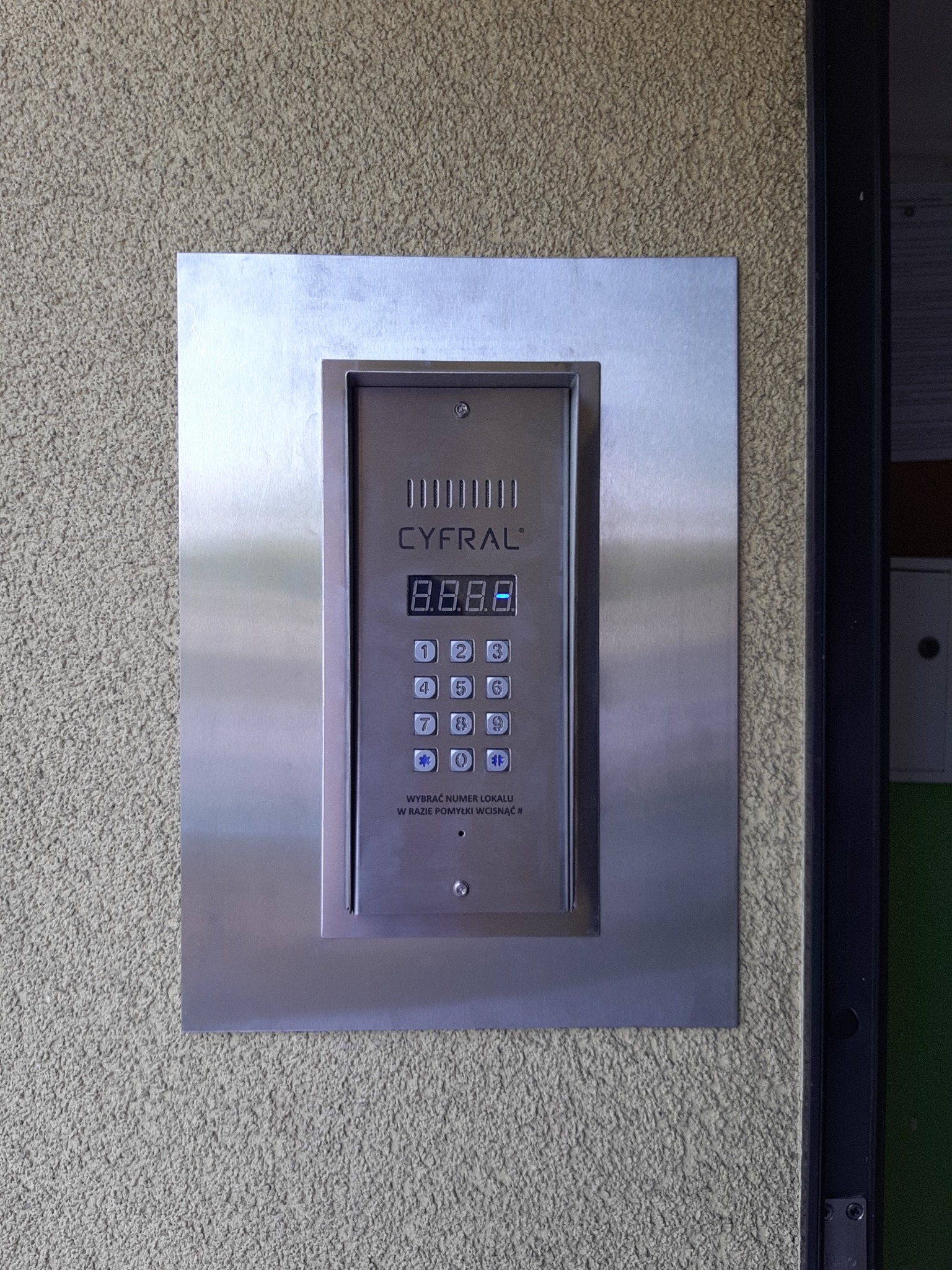 Panel domofonowy Cyfral z metalową obudową zamontowany na chropowatej ścianie budynku.