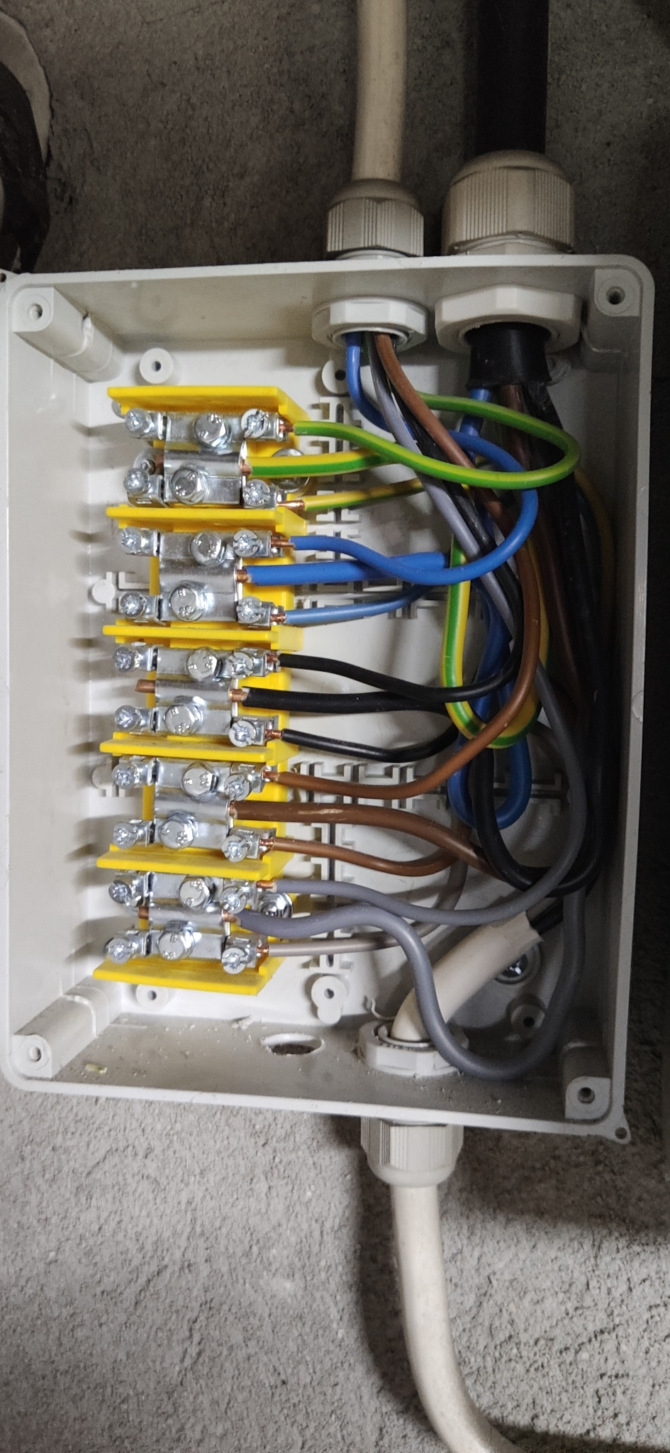 Rozdzielnica elektryczna z żółtymi listwami zaciskowymi, widoczne przewody w różnych kolorach, w tym brązowy, niebieski, czarny, zielono-żółty, szary i biały, w szarej obudowie na tle szarej ściany.