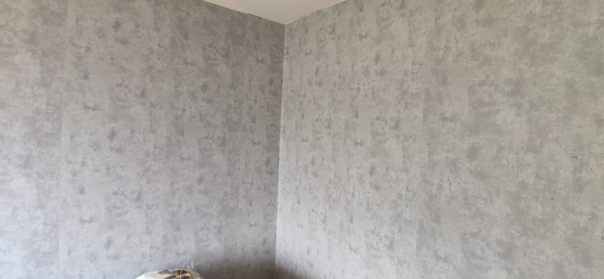 Fragment rogu pokoju z szarą, teksturowaną tapetą na ścianach, ukazujący wykończenie wnętrza.