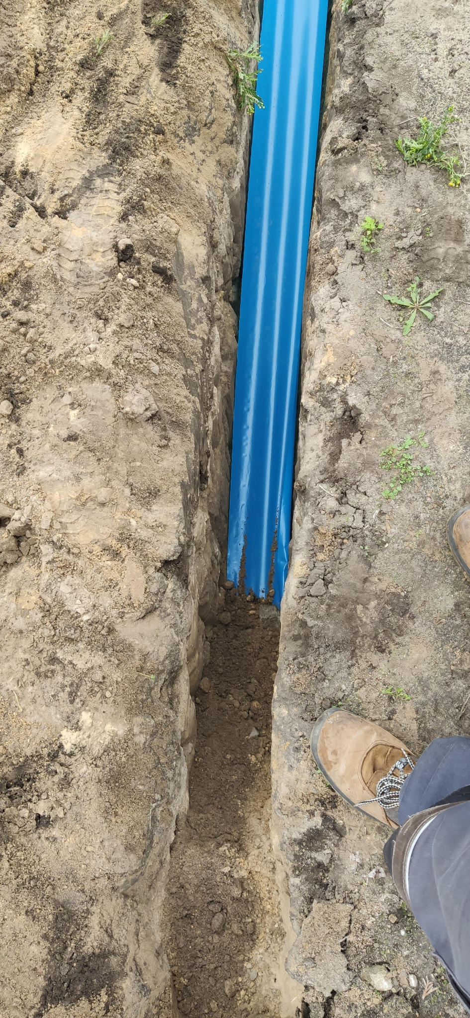 Niebieska rura w wykopie ziemnym, widoczny fragment buta roboczego
