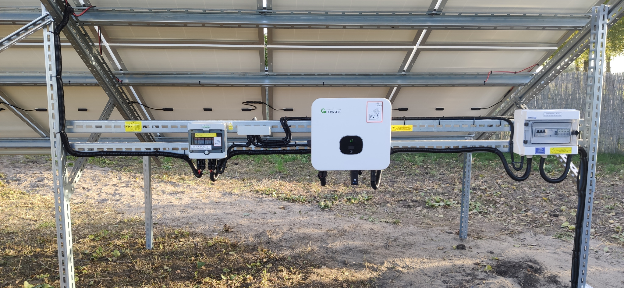 Widok od spodu na instalację paneli fotowoltaicznych z widocznym inwerterem Growatt, skrzynką przyłączeniową i okablowaniem, zamontowanych na metalowej konstrukcji na gruncie.