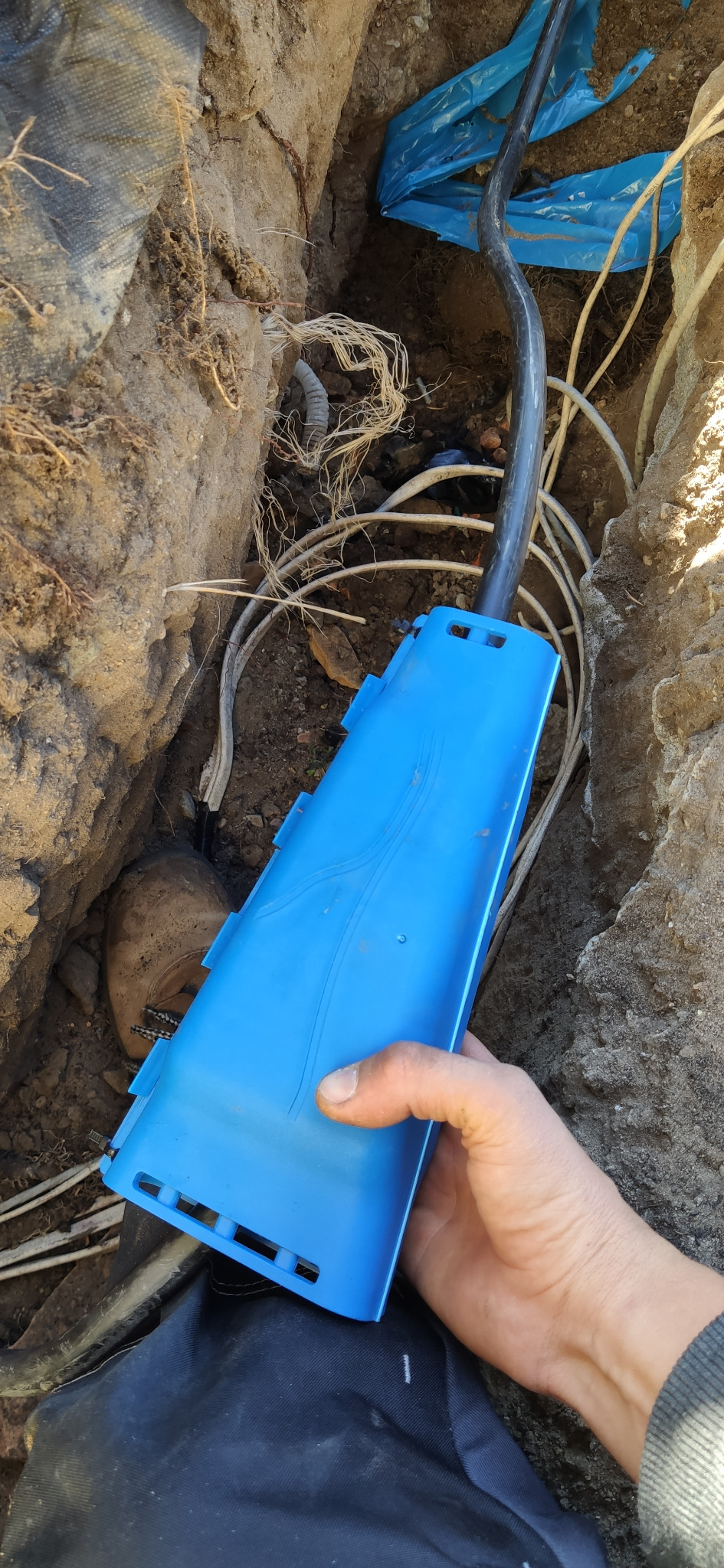 Ręka trzyma niebieską skrzynkę przyłączeniową kabli w wykopie ziemnym z widocznymi kablami i rurami osłonowymi.