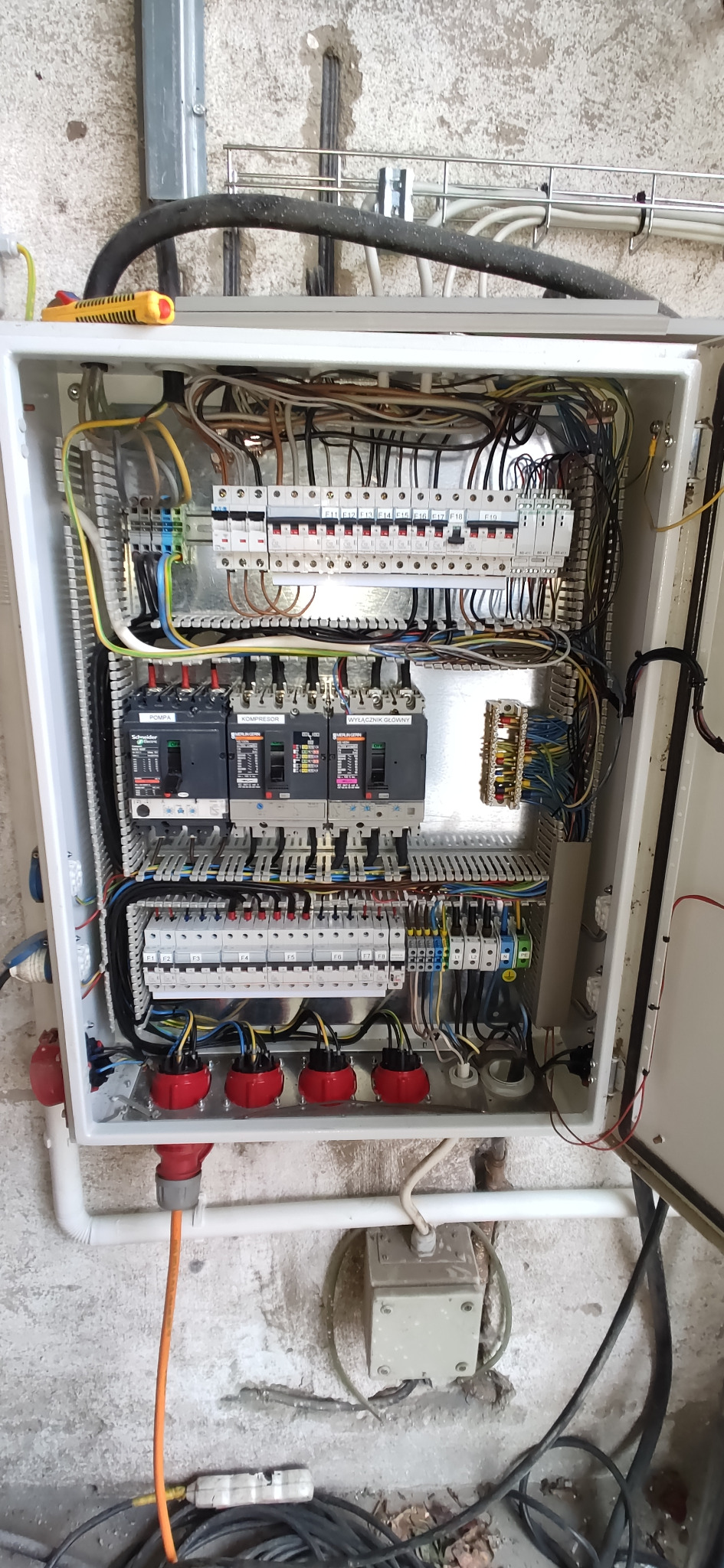 Otwarta skrzynka elektryczna z bezpiecznikami, okablowaniem i gniazdami wtykowymi, widoczne oznaczenia 'Pompa', 'Kompresor' i 'Wyłącznik Główny' na poszczególnych wyłącznikach.