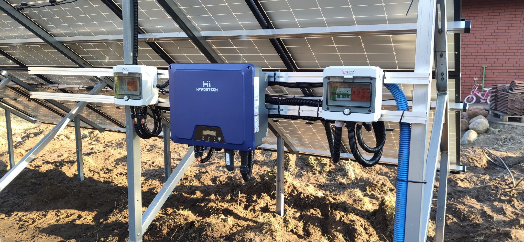 Instalacja paneli fotowoltaicznych w Ciechocinku, widoczny niebieski inwerter Hypontech i skrzynki przyłączeniowe na konstrukcji wsporczej, w tle ceglany budynek.