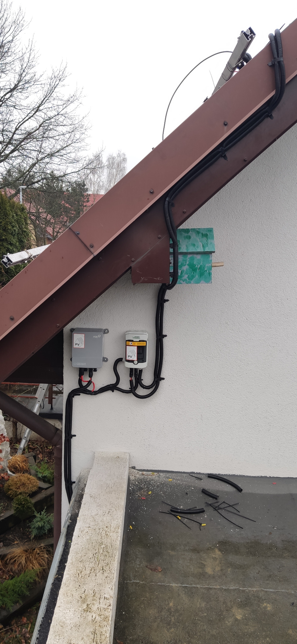 Dwa falowniki fotowoltaiczne zamontowane na elewacji budynku pod okapem dachu, widoczne kable w peszlach i złączki.