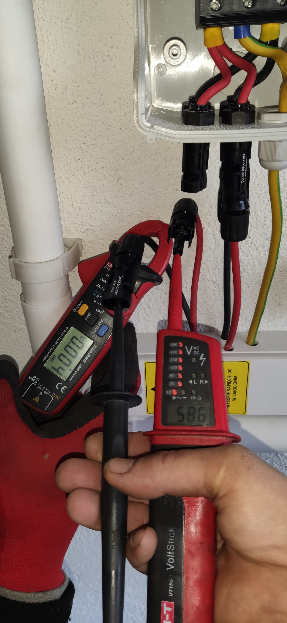 Pomiar napięcia instalacji elektrycznej za pomocą multimetru cęgowego i wskaźnika napięcia VoltStick, widoczne przewody w skrzynce przyłączeniowej.