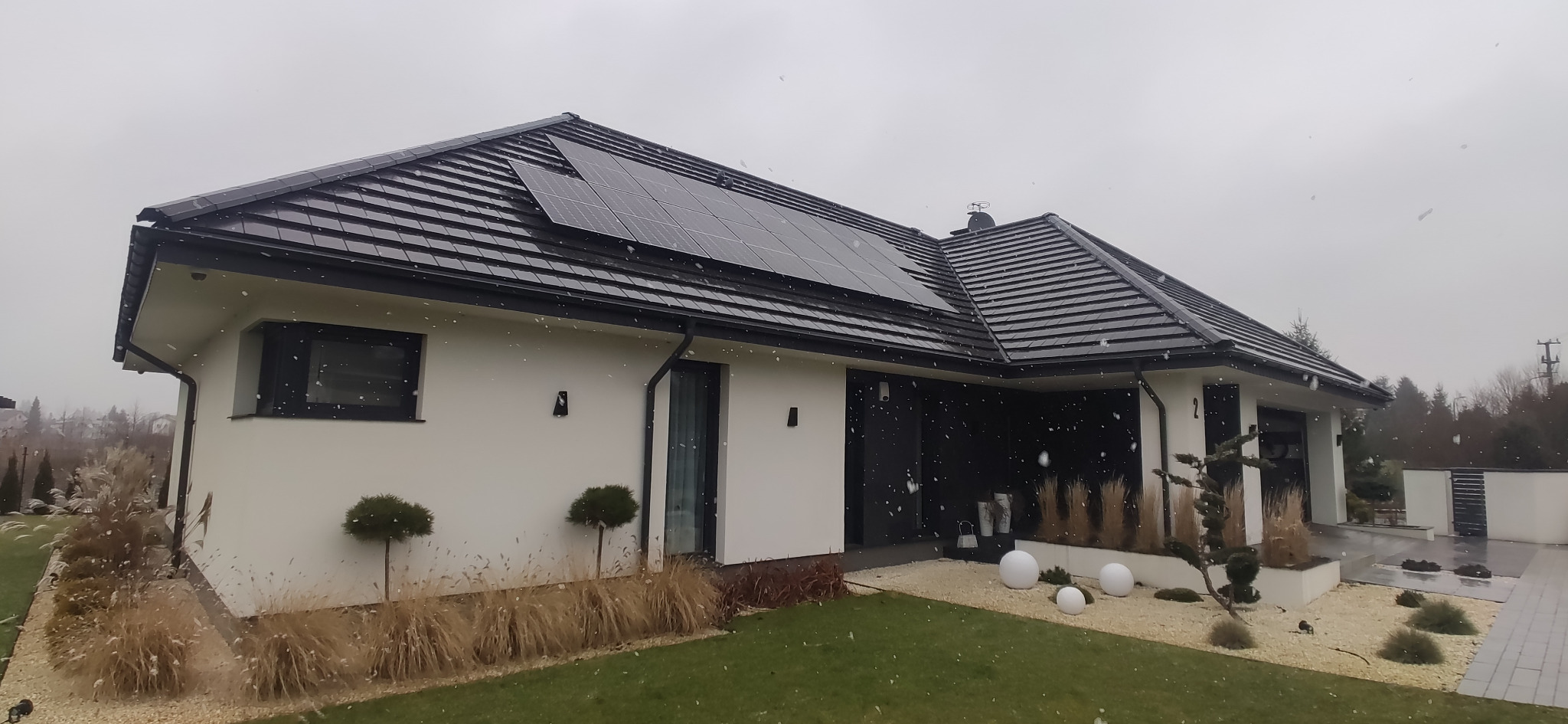 Nowoczesny dom jednorodzinny z panelami słonecznymi na dachu, widok z ogrodu podczas opadów śniegu.