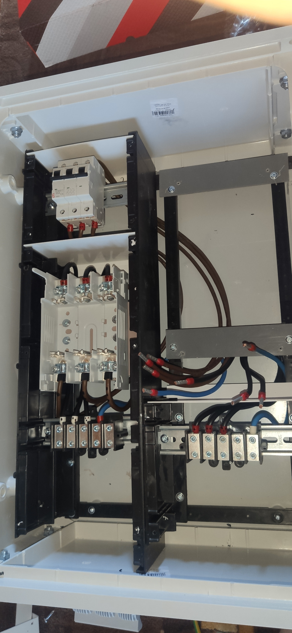 Wnętrze skrzynki elektrycznej z zamontowanymi bezpiecznikami, przewodami i złączkami, widoczne brązowe i niebieskie przewody z czerwonymi końcówkami.