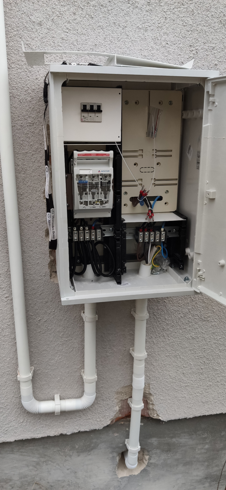 Otwarta skrzynka elektryczna na zewnątrz budynku z widocznym licznikiem, bezpiecznikami i przewodami, zamontowana na szarej ścianie z rurami PCV.