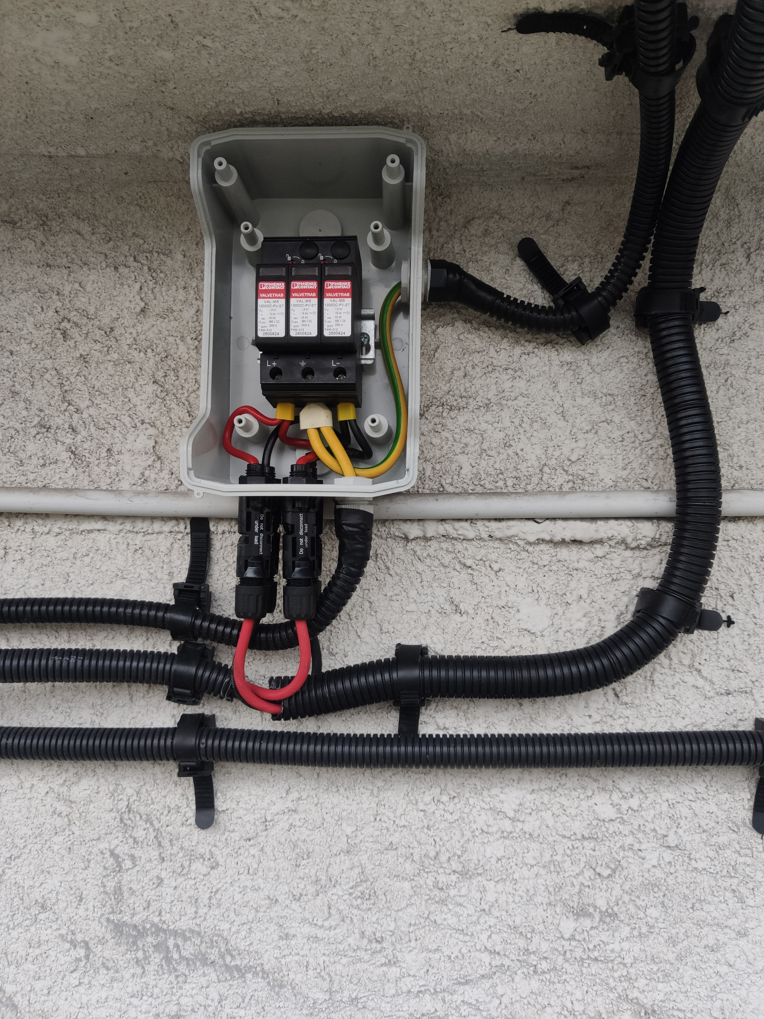 Instalacja elektryczna zabezpieczająca przed przepięciami w szarej skrzynce na teksturowanej ścianie, z widocznymi przewodami w czarnych peszlach.