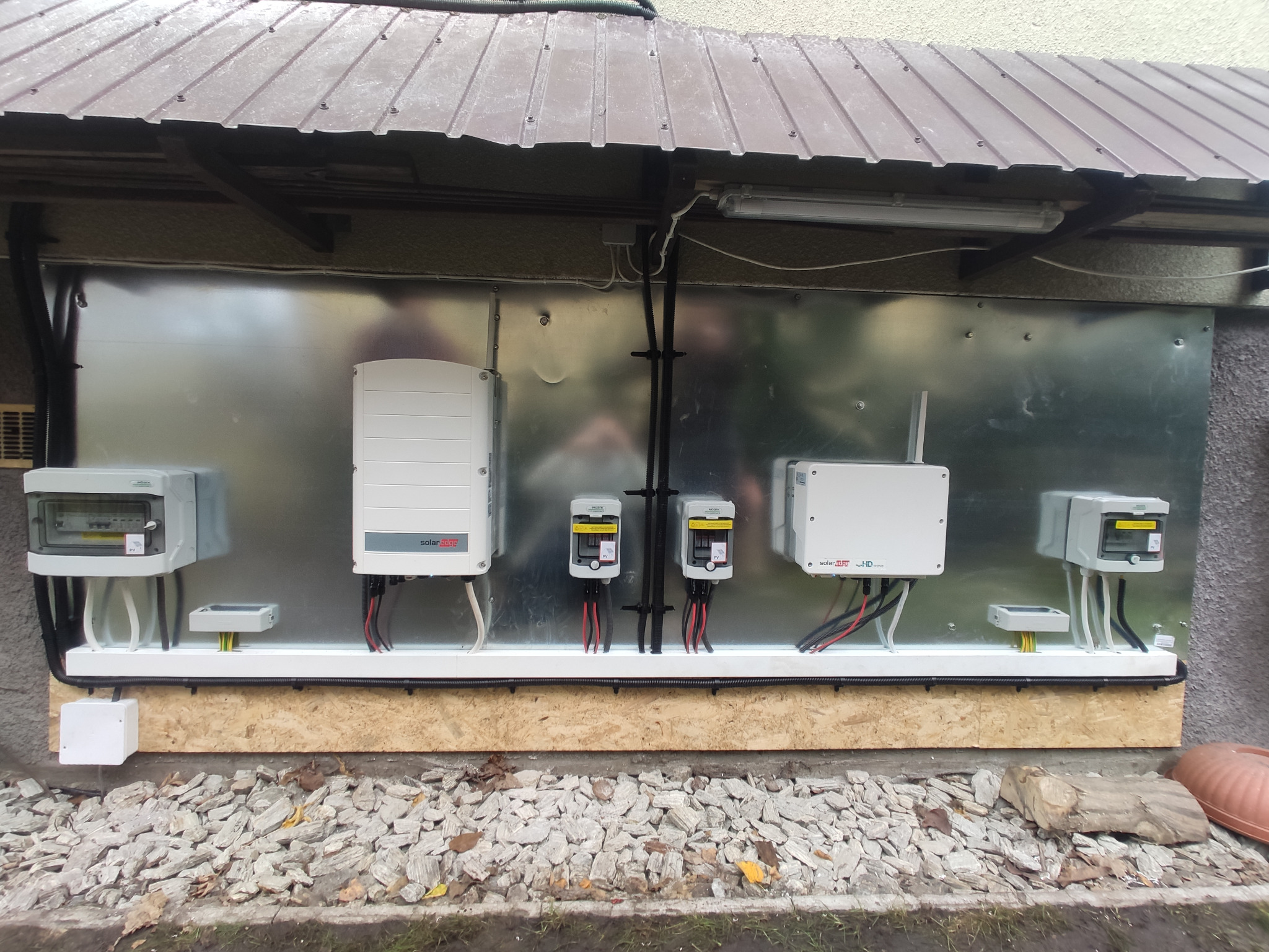 Zewnętrzna instalacja elektryczna pompy ciepła z falownikami SolarEdge i zabezpieczeniami na srebrnej blasze, widok z poziomu gruntu na tle kamieni i elewacji budynku z brązowym zadaszeniem.