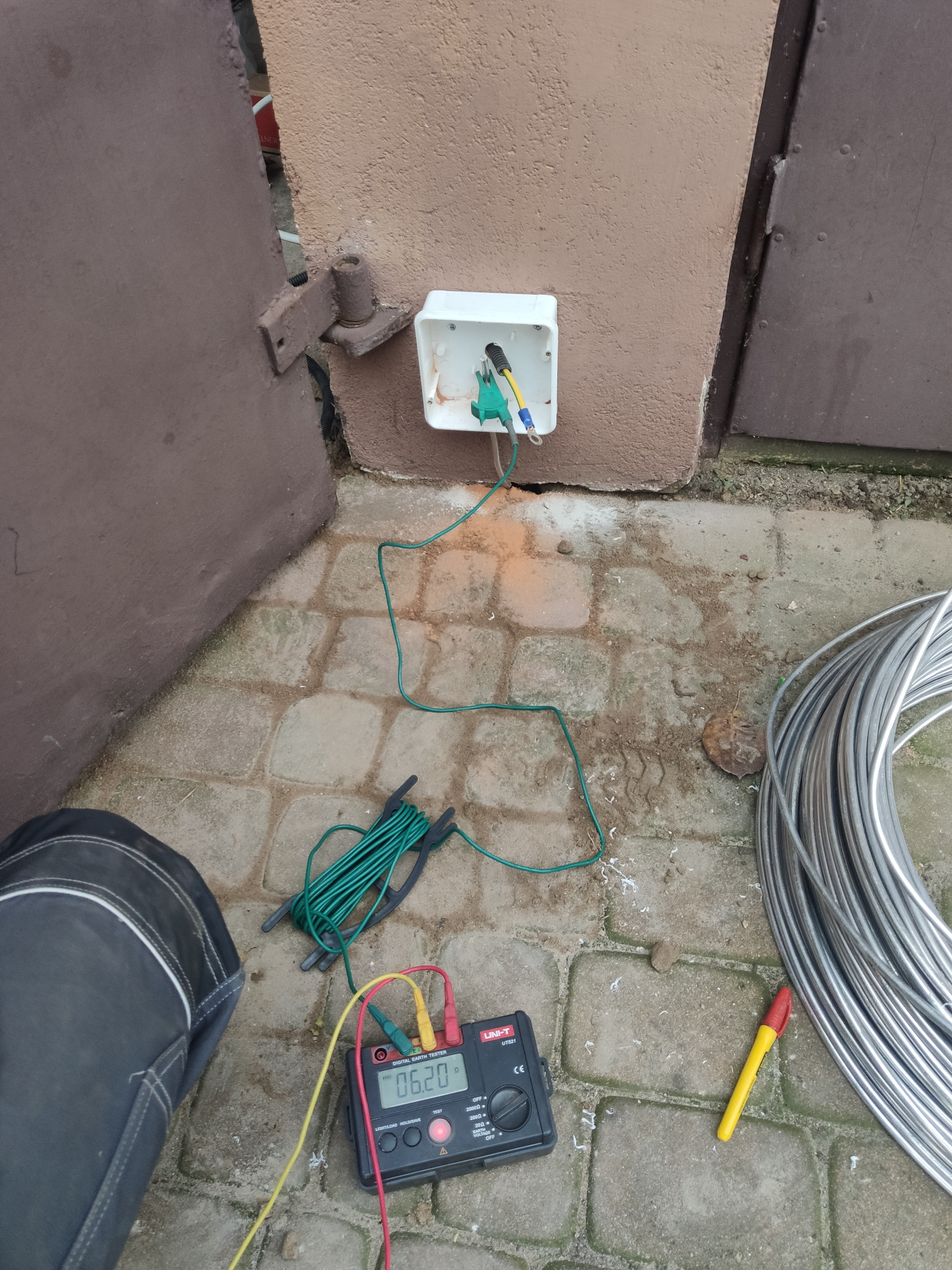 Pomiar uziemienia przy użyciu miernika cyfrowego UNI-T UT821, z widoczną skrzynką elektryczną na elewacji i wiązką kabli uziemiających na brukowanym podłożu.