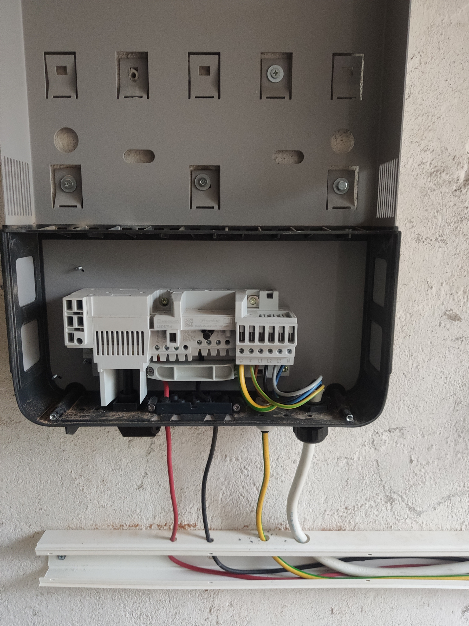 Otwarta szara skrzynka elektryczna z widocznymi przewodami w kolorach żółtym, zielonym, czarnym i czerwonym, przymocowana do szorstkiej, białej ściany.