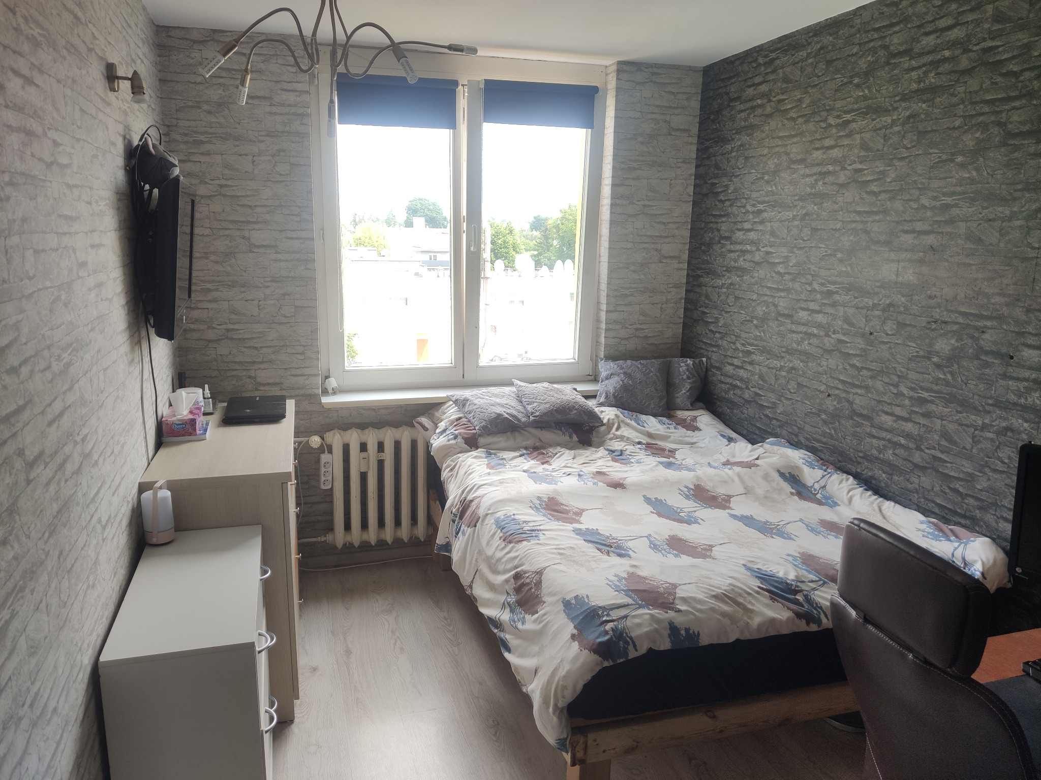 Pokój z łóżkiem, grzejnikiem pod oknem i ścianą imitującą kamień, widok na zewnątrz przez okno.