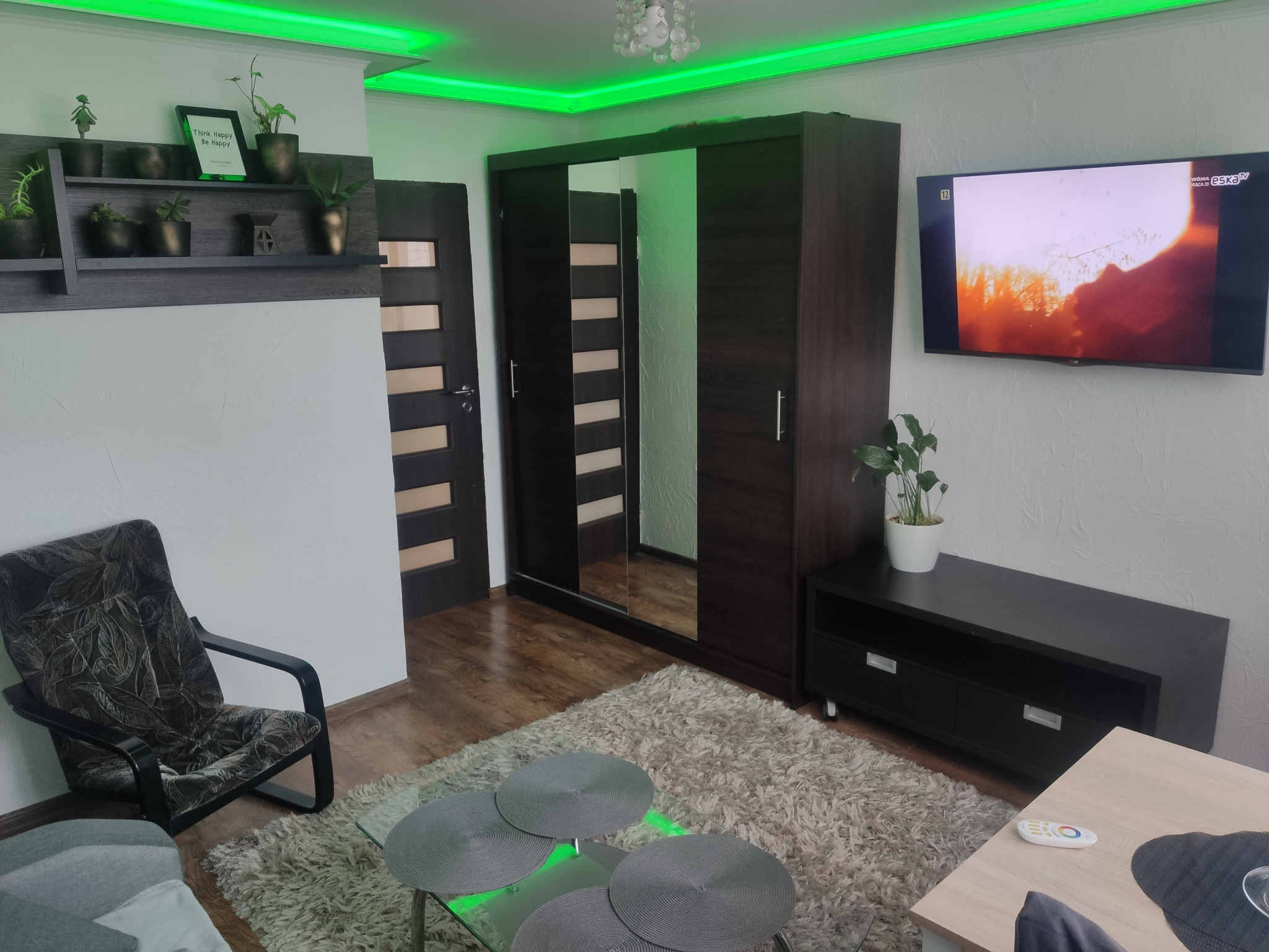 Jasne wnętrze pokoju z zielonym oświetleniem LED, ciemnymi meblami, szarą wykładziną i telewizorem z włączonym programem.