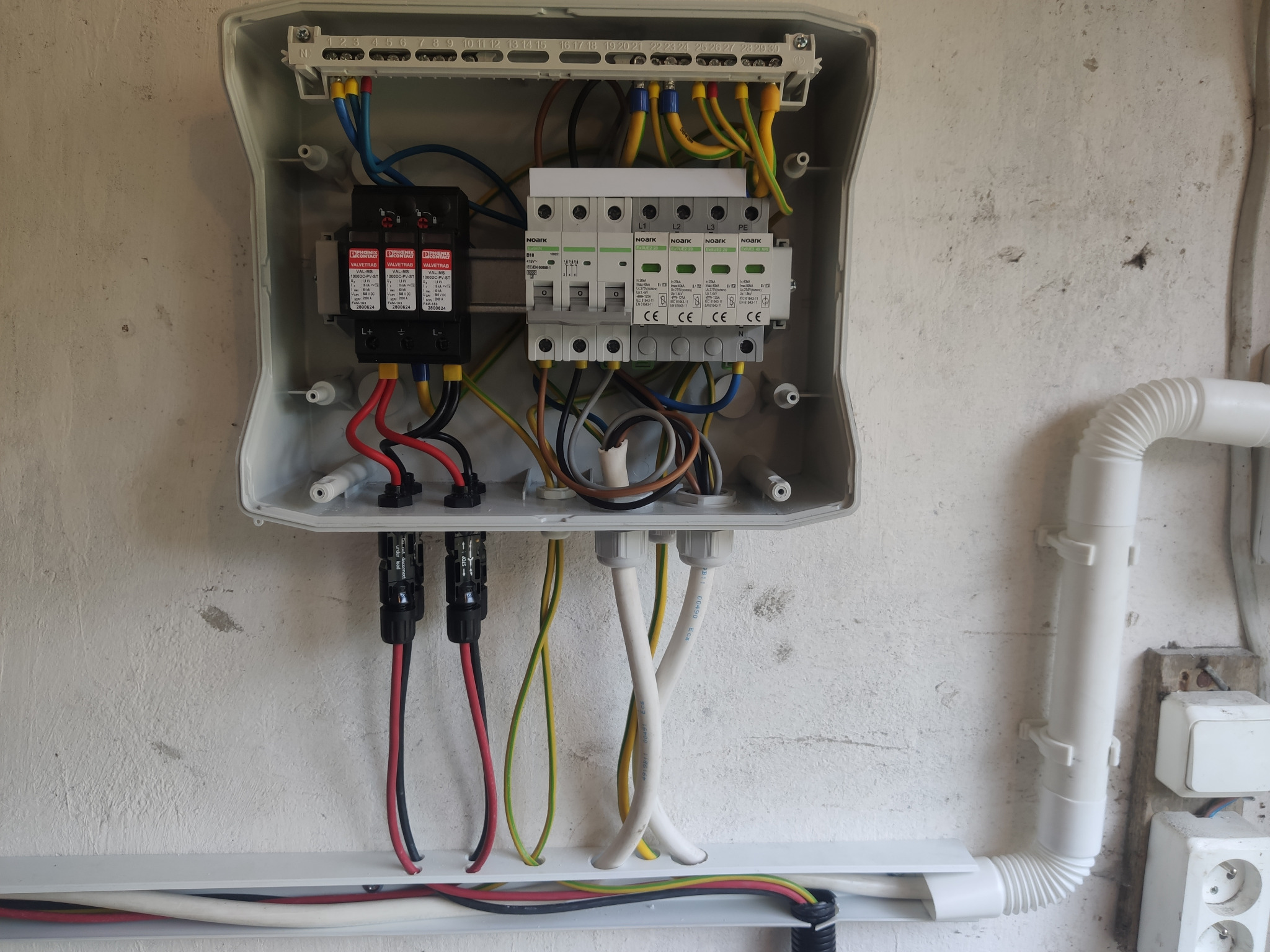 Rozdzielnica elektryczna z zabezpieczeniami przepięciowymi i wyłącznikami, widoczne kable i przewody w kolorach, zamontowana na szarej ścianie.