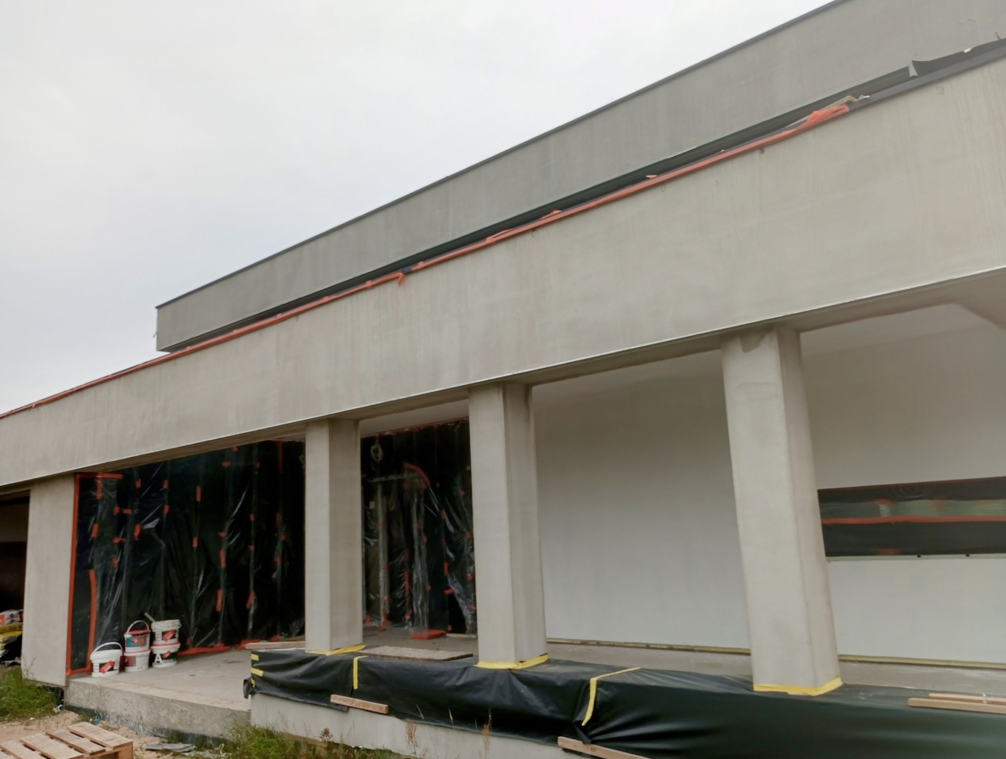 Budynek w trakcie przygotowania do malowania elewacji, okna zabezpieczone folią, widoczne puszki z farbą i paleta drewniana.