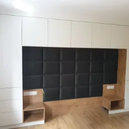 Biała zabudowa meblowa z szafkami nad łóżkiem, czarny panel tapicerowany w kwadraty i drewniane szafki nocne z gniazdkami.