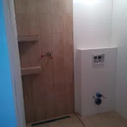 Wykończona łazienka z panelami imitującymi drewno na ścianie prysznicowej i podłodze, białe płytki na ścianie z zabudowaną spłuczką WC i widocznymi przyłączami hydraulicznymi.