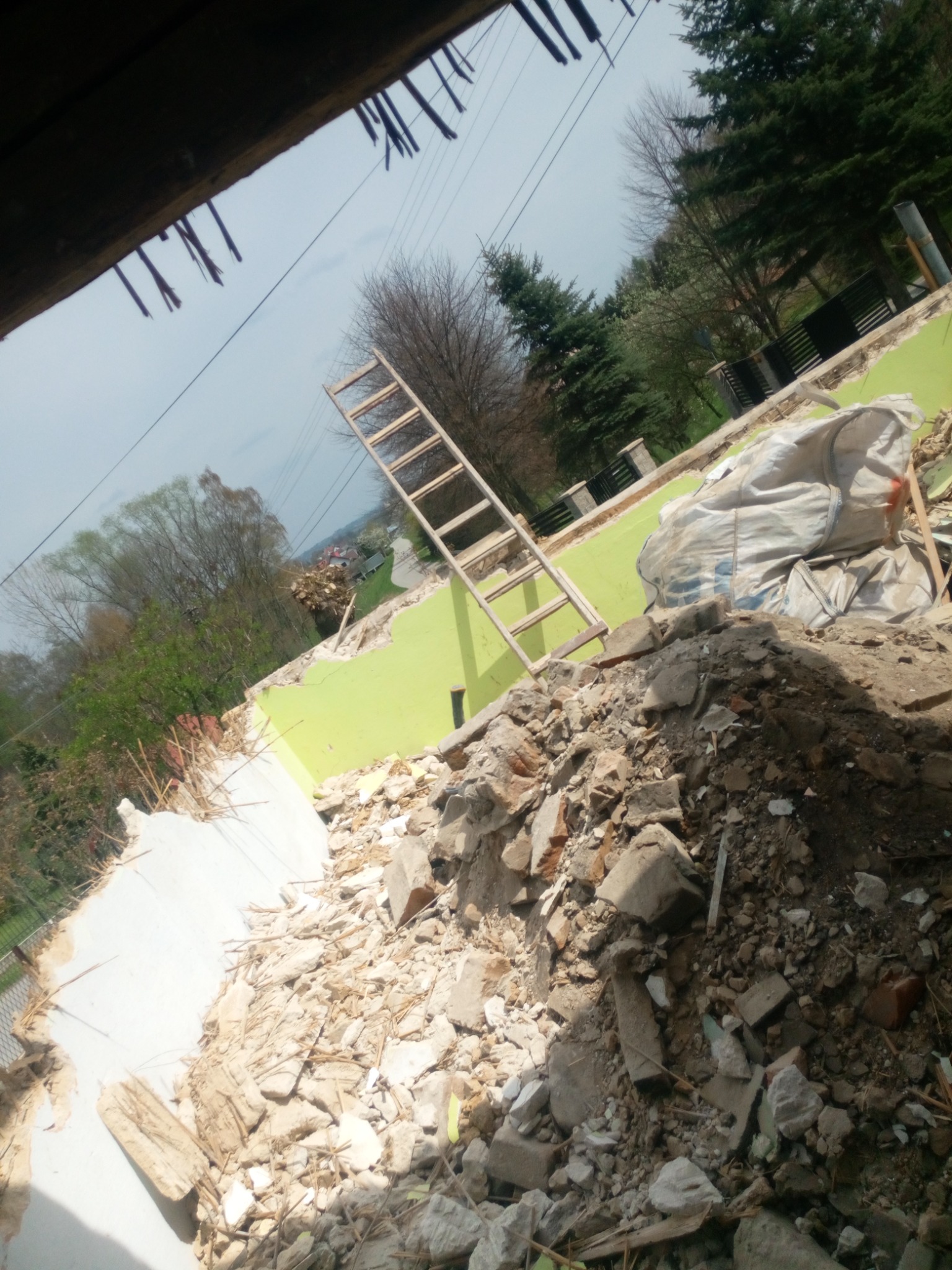 Fragment zdemontowanej ściany z pozostałościami zielonej farby, sterta gruzu, drewniana drabina oparta o ścianę, widok z góry na teren rozbiórki budynku.