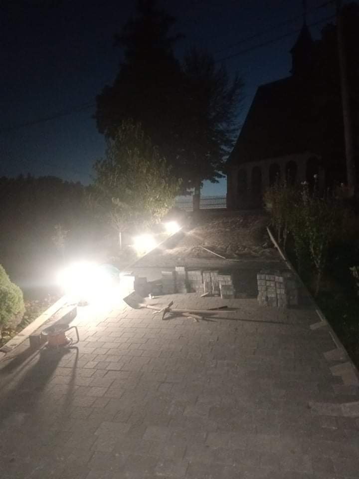 Układanie kostki brukowej na podjeździe w wieczornym oświetleniu, widoczne ułożone kostki, narzędzia i fragment kapliczki w tle.