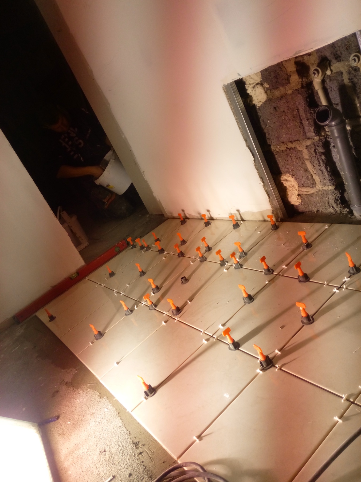 Układanie jasnych płytek podłogowych z użyciem pomarańczowych klipsów poziomujących, widoczna poziomica i fragment instalacji hydraulicznej przy ścianie.