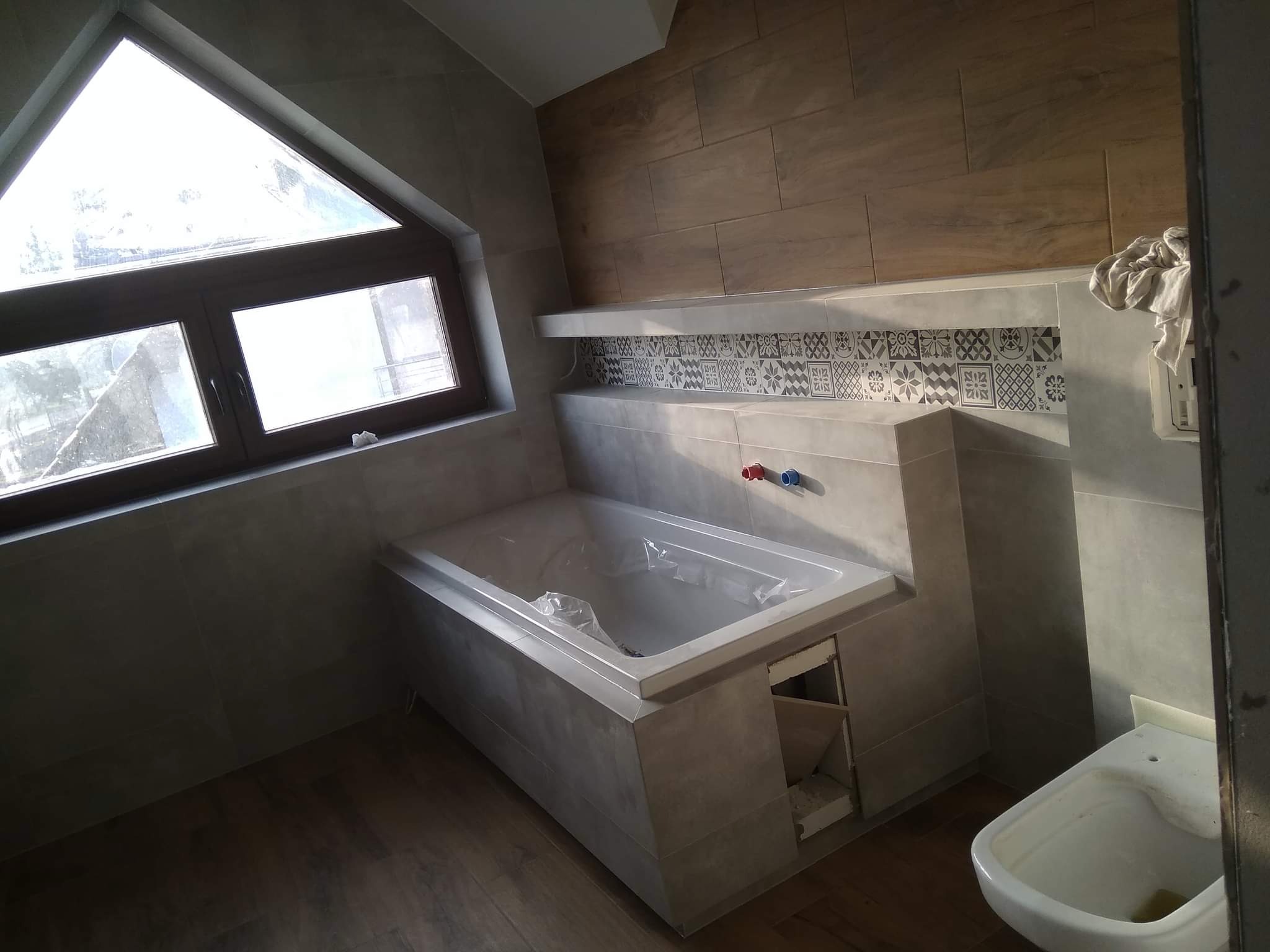 Wnętrze łazienki w trakcie wykańczania: obudowana wanna z widocznymi instalacjami, okno dachowe trójkątne, płytki imitujące drewno na ścianie, dekoracyjny pas płytek nad wanną.