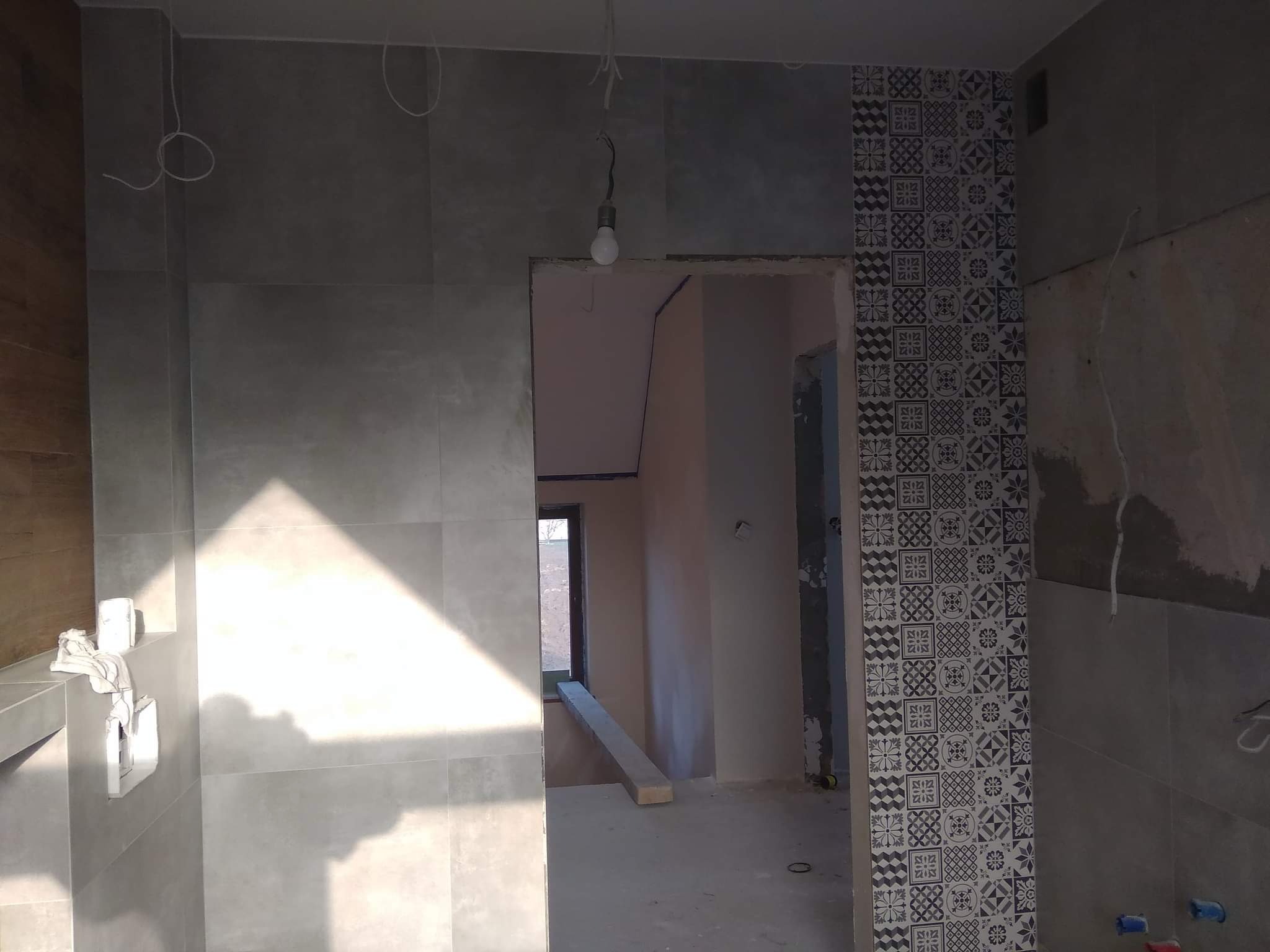 Wnętrze łazienki w trakcie remontu z szarymi płytkami ściennymi i dekoracyjną kolumną z płytek wzorzystych, widoczne przewody elektryczne i surowa żarówka.