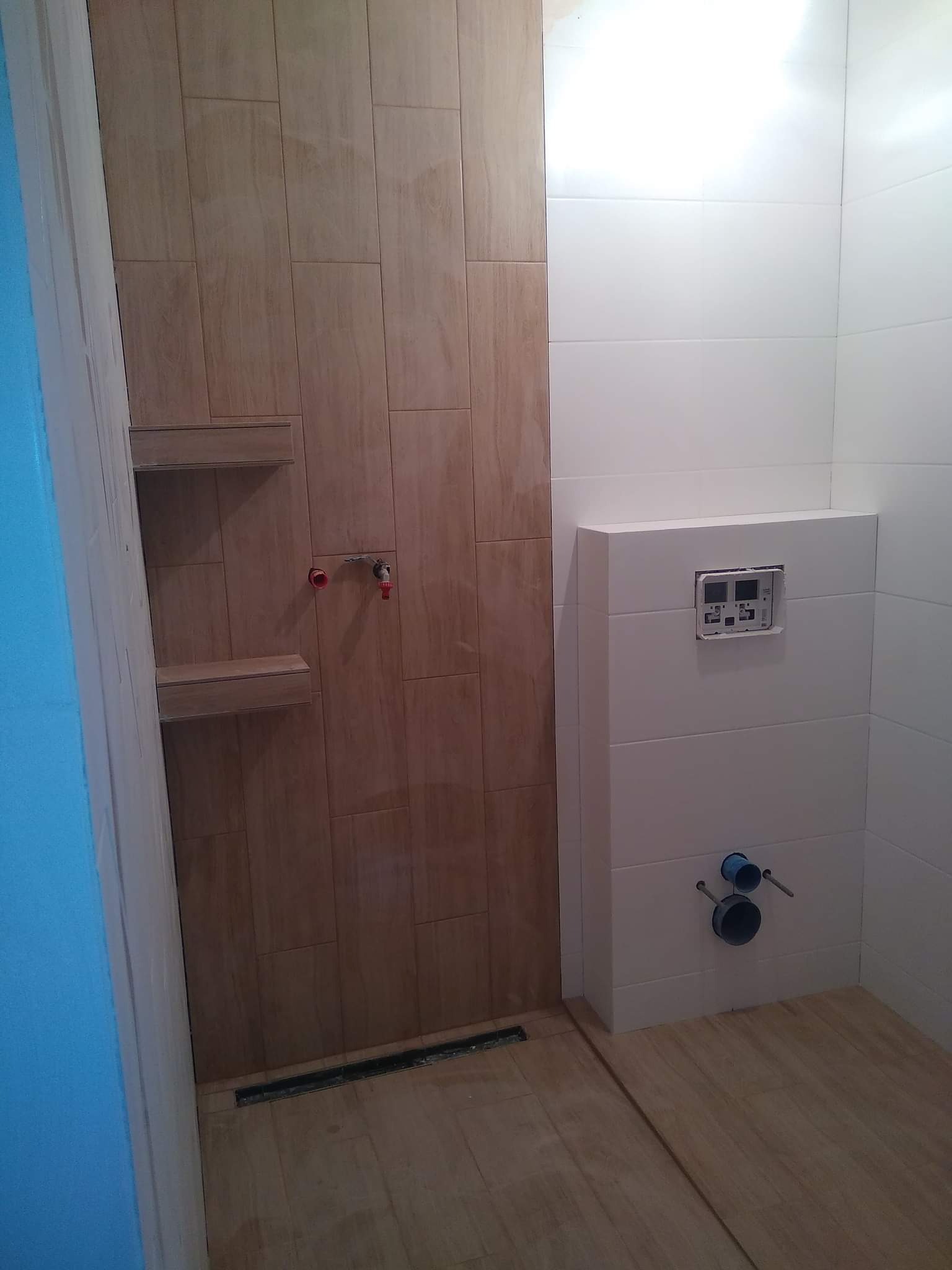 Wykończona łazienka z panelami imitującymi drewno na ścianie prysznicowej i podłodze, białe płytki na ścianie z zabudowaną spłuczką WC i widocznymi przyłączami hydraulicznymi.