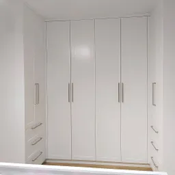 Biała szafa wnękowa z czterema drzwiami i szufladami po bokach, z długimi, prostymi uchwytami, dopasowana do przestrzeni od podłogi do sufitu.