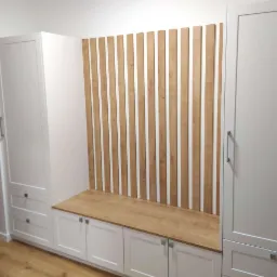 Biała zabudowa z szafami i siedziskiem z drewnianym blatem, dekoracyjna ścianka z pionowych listew z jasnego drewna.