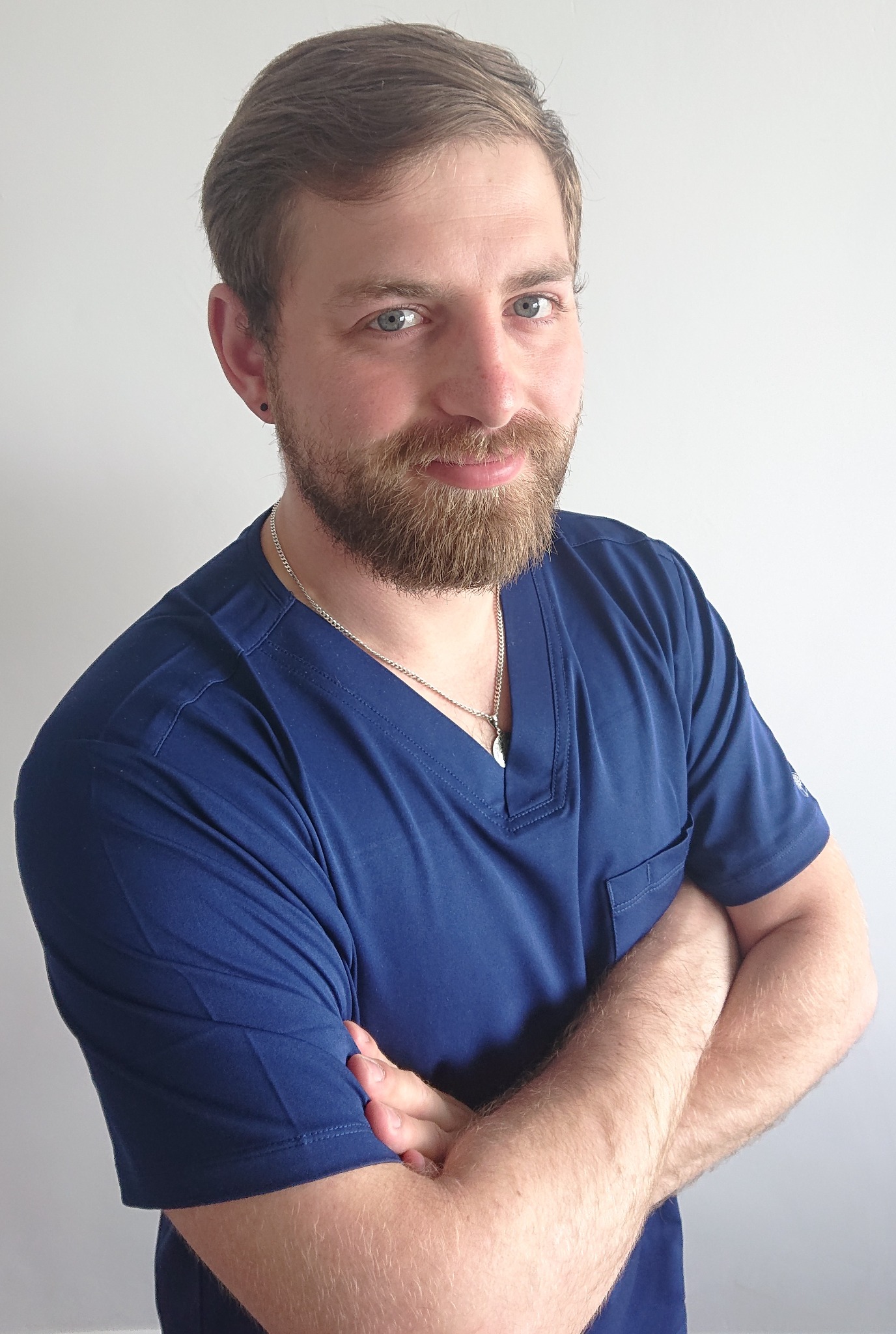 Uśmiechnięty mężczyzna w niebieskim uniformie medycznym, ze skrzyżowanymi rękami i srebrnym łańcuszkiem, pozuje na tle jasnej ściany.