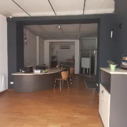 Wnętrze biura z recepcją, drewnianą podłogą, grzejnikiem przy białej ścianie, szarą ścianą z szafką i obrazem, widok na dalsze pomieszczenie z biurkami i krzesłami.