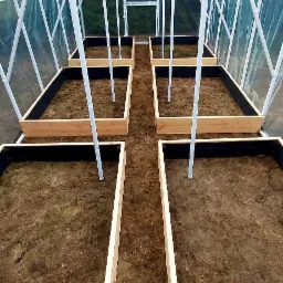 Wnętrze szklarni z drewnianymi podwyższonymi grządkami wyłożonymi czarną folią, przygotowanymi pod uprawę roślin.