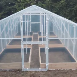 Nowo postawiona, foliowa szklarnia z drewnianymi podwyższonymi grządkami wypełnionymi ziemią, widok z zewnątrz na tle zieleni.